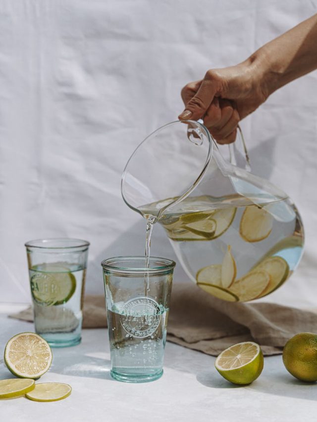 Crop person holding jug and pouring fresh lemonade into glass on white textile background